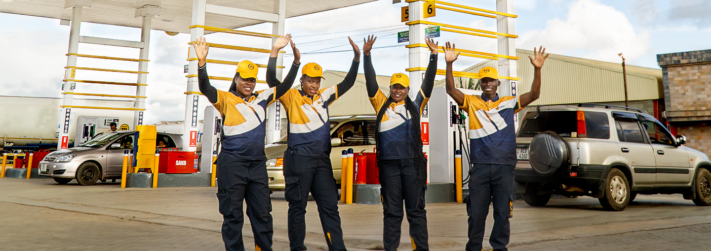 Four staff members dressed in company uniform with their arms raised and smiling, standing on the forecourt of an UNO Energies filling station