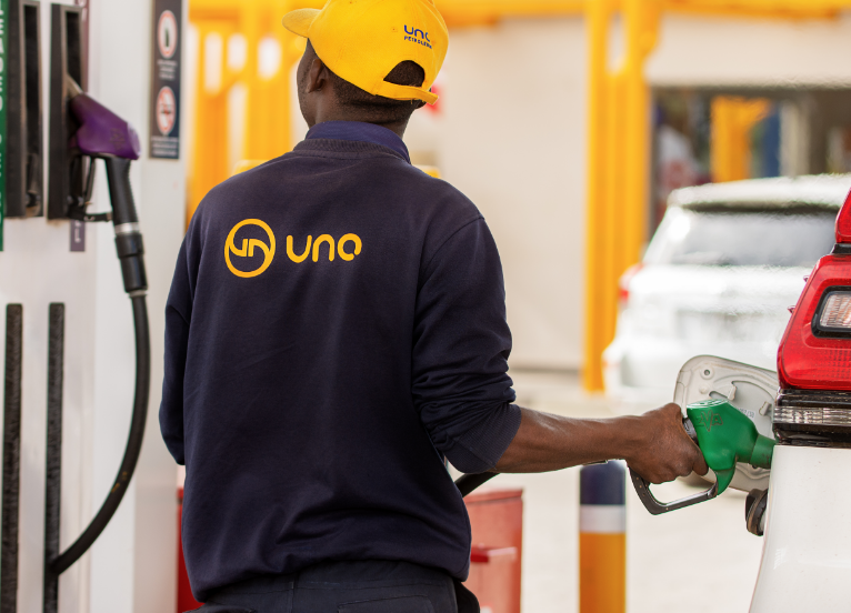 An UNO Energies employee in a branded blue shirt and yellow cap with his back to the camera, adding fuel to a car and watching the readout screen on the fuel pump